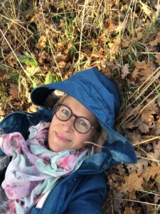 Julia Falkenstein liegt lächelnd auf dem Waldboden eines sonnigen Mischwaldes. Das Bild symbolisiert die Vielfalt und Balance des Darmmikrobioms.
